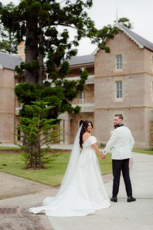 Bride and groom hold hands walking away from a historic sandstone building, bride wearing a long cathedral veil.