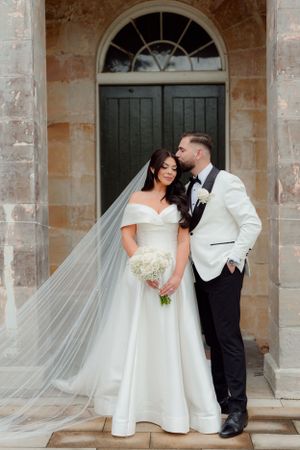 Bride in off-shoulder gown with long veil and groom in white tuxedo before stone doorway.
