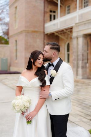 Bride in off-shoulder white gown and groom in white tuxedo embrace tenderly outside a historic sandstone venue.