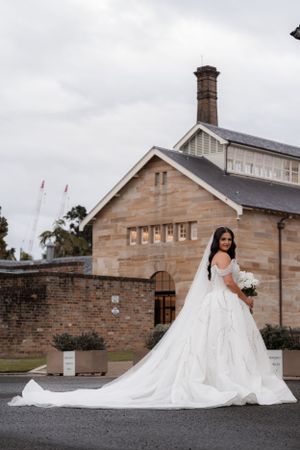 Bride in elegant white ball gown with long train poses outside historic sandstone building on her wedding day.