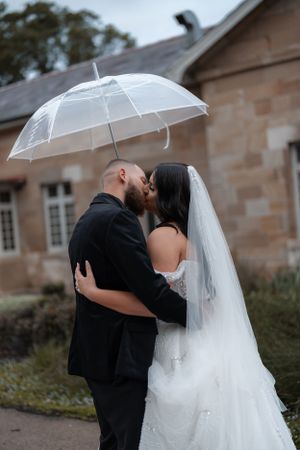 Bride and groom share a romantic kiss under a clear umbrella in the rain outside a historic stone building.