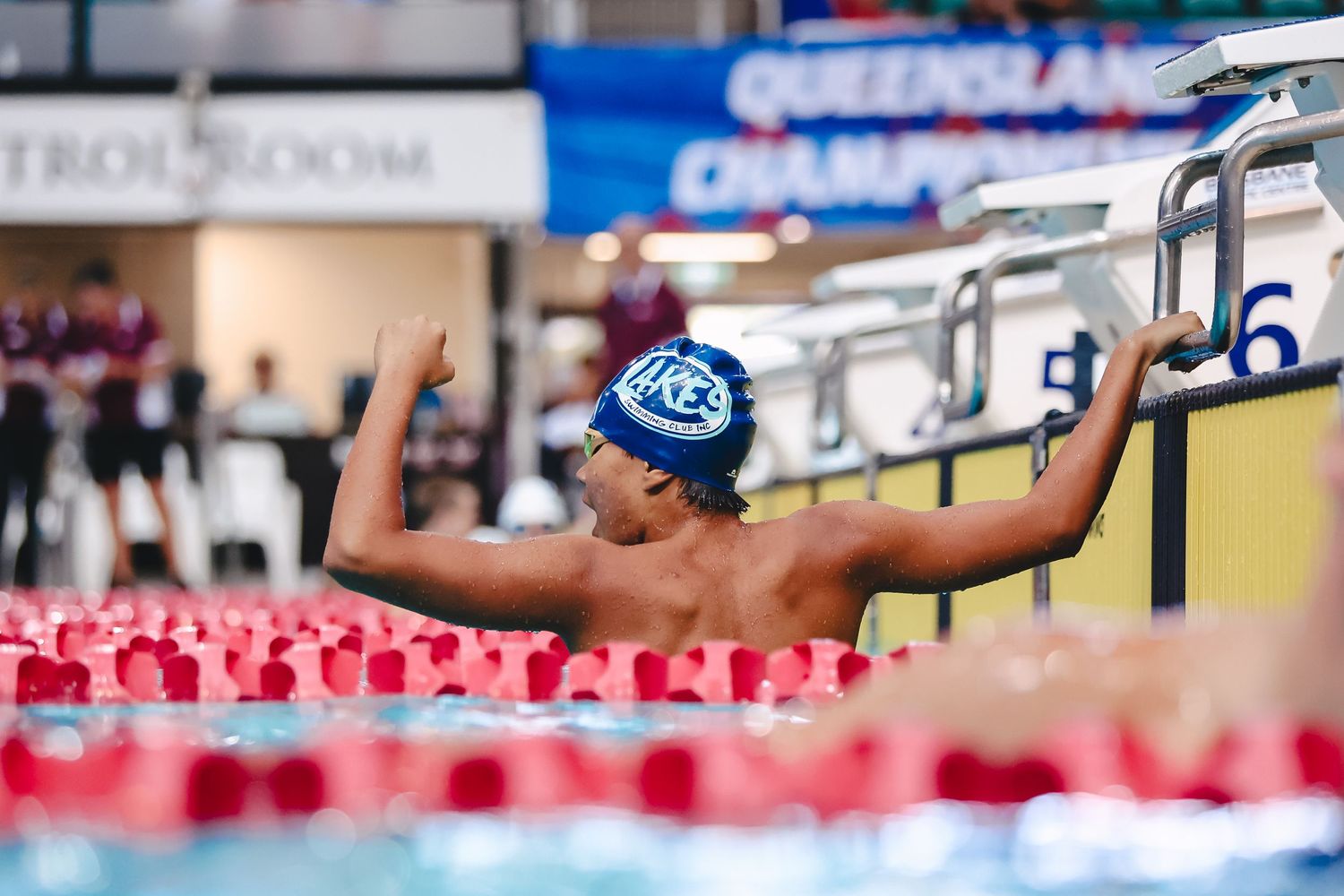Swimmer in a blue cap celebrates by raising both arms at the side of a swimming lane surrounded by red lane markers.