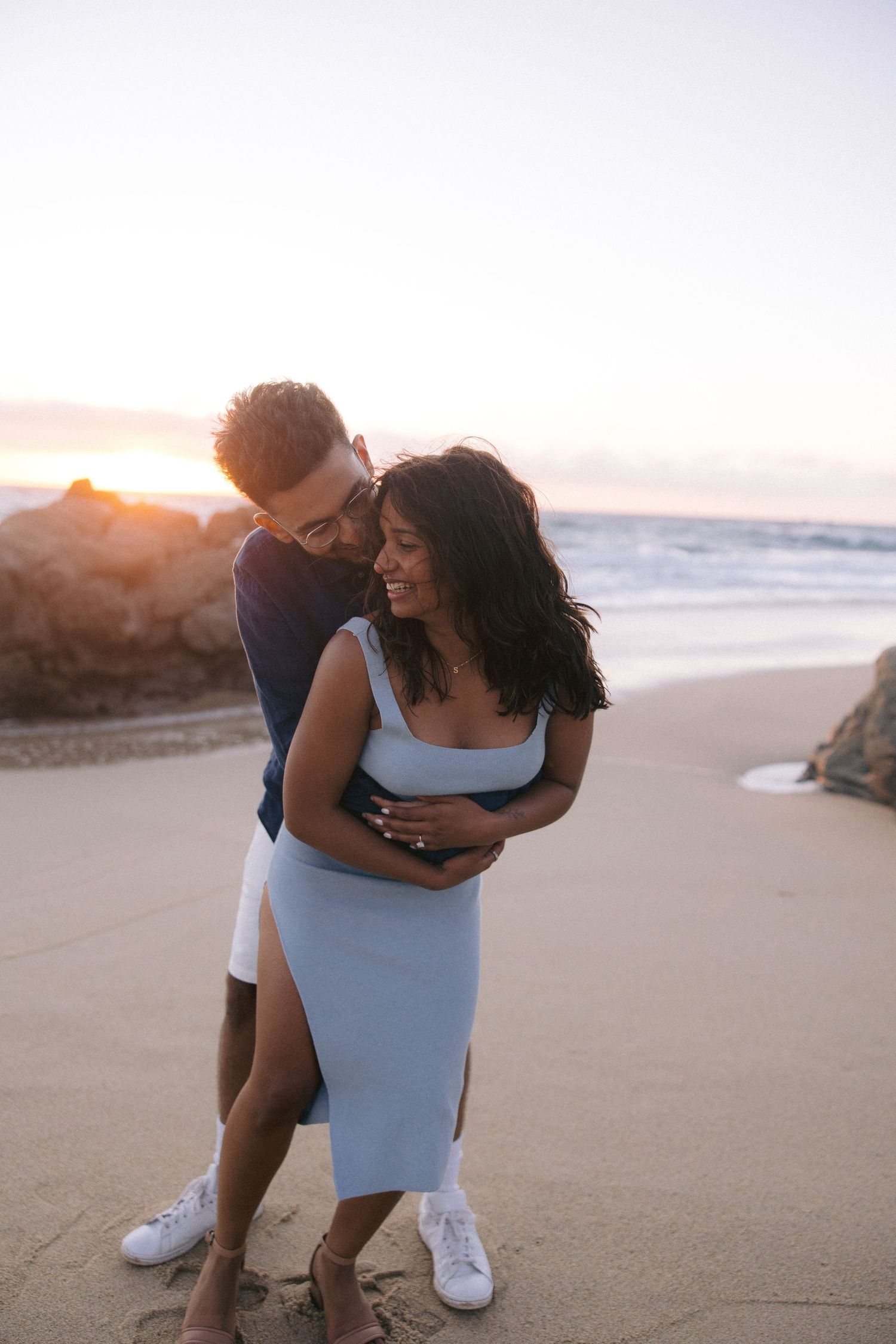A couple embraces and laughs together on a sandy beach at sunset with ocean waves and rocks in the background.