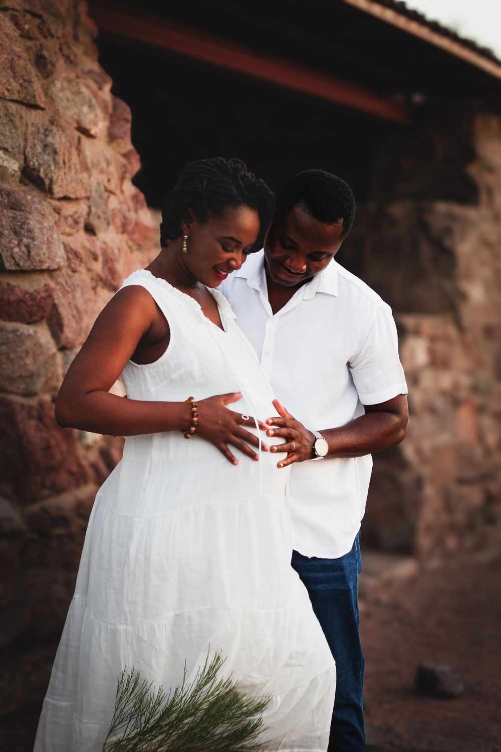 Couple in white clothing shares intimate moment during pregnancy photoshoot against stone wall.