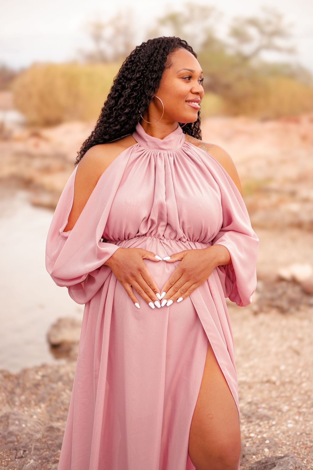 Expectant mother in flowing pink maxi dress poses outdoors in desert setting during maternity photoshoot.