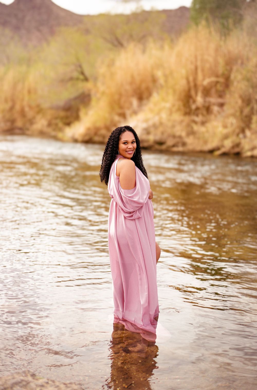 A person in a flowing pink dress stands in a shallow river with mountains and brush in the background during sunset.