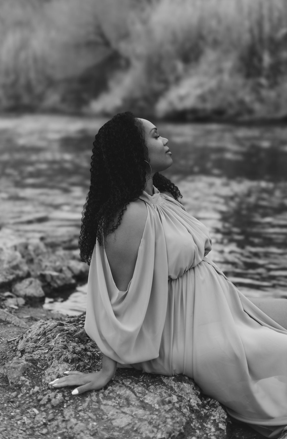 A person in a flowing white dress poses gracefully on a rock by a riverbank in a black and white photograph.