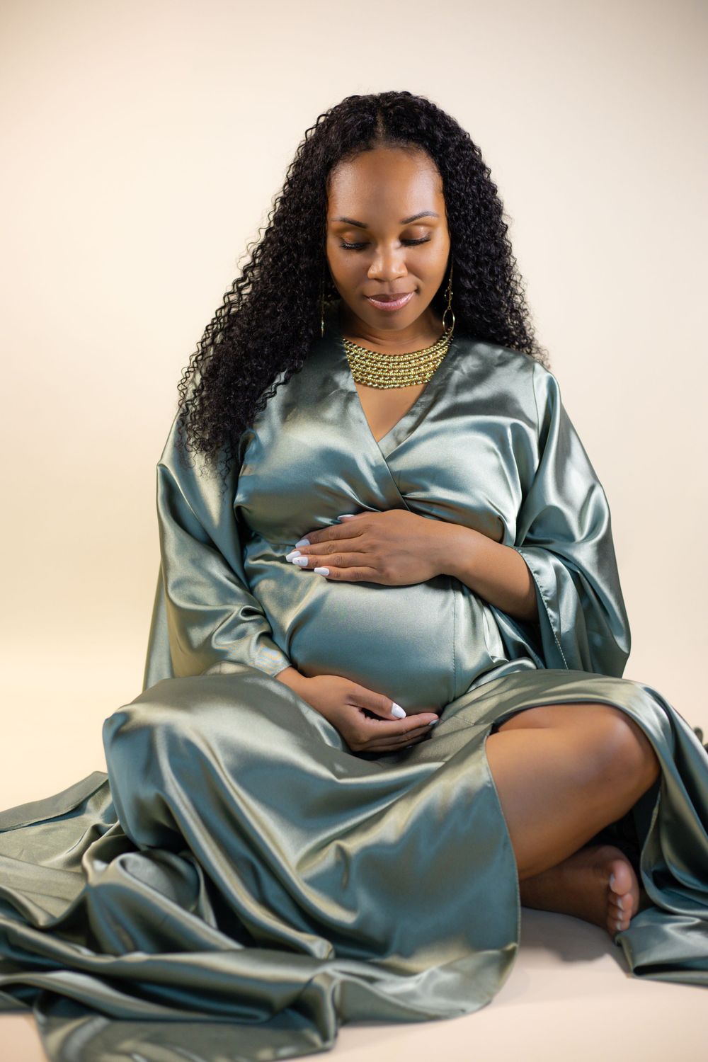 Beautiful maternity photo with expecting mother in flowing teal silk robe and gold necklace against soft beige backdrop.