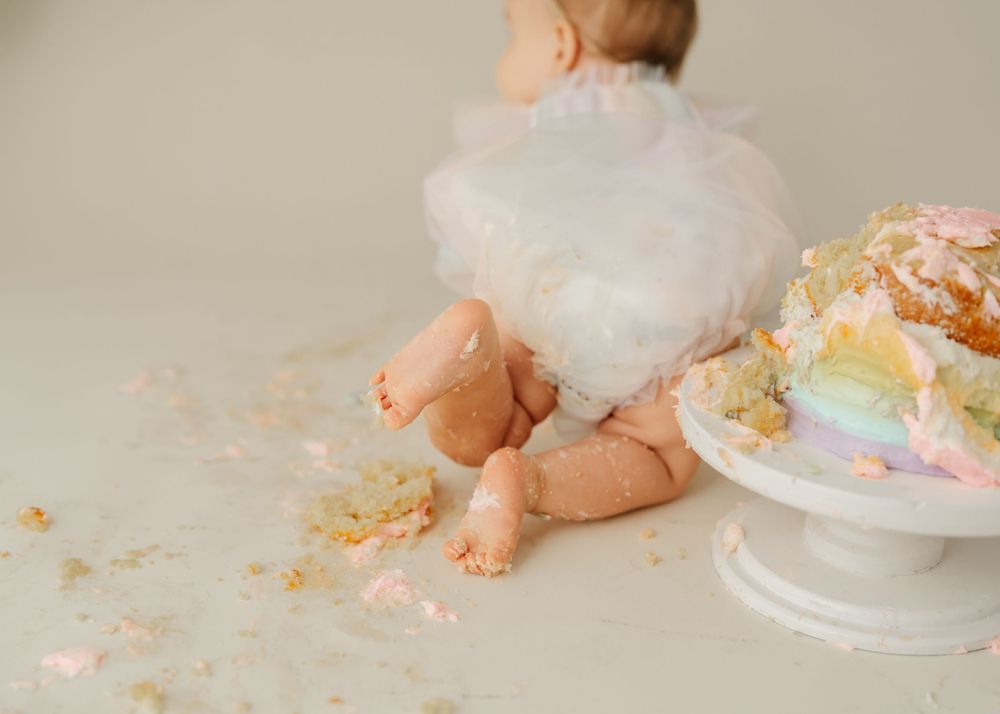 Small feet and legs visible next to a pastel rainbow cake with crumbs scattered on a light floor.