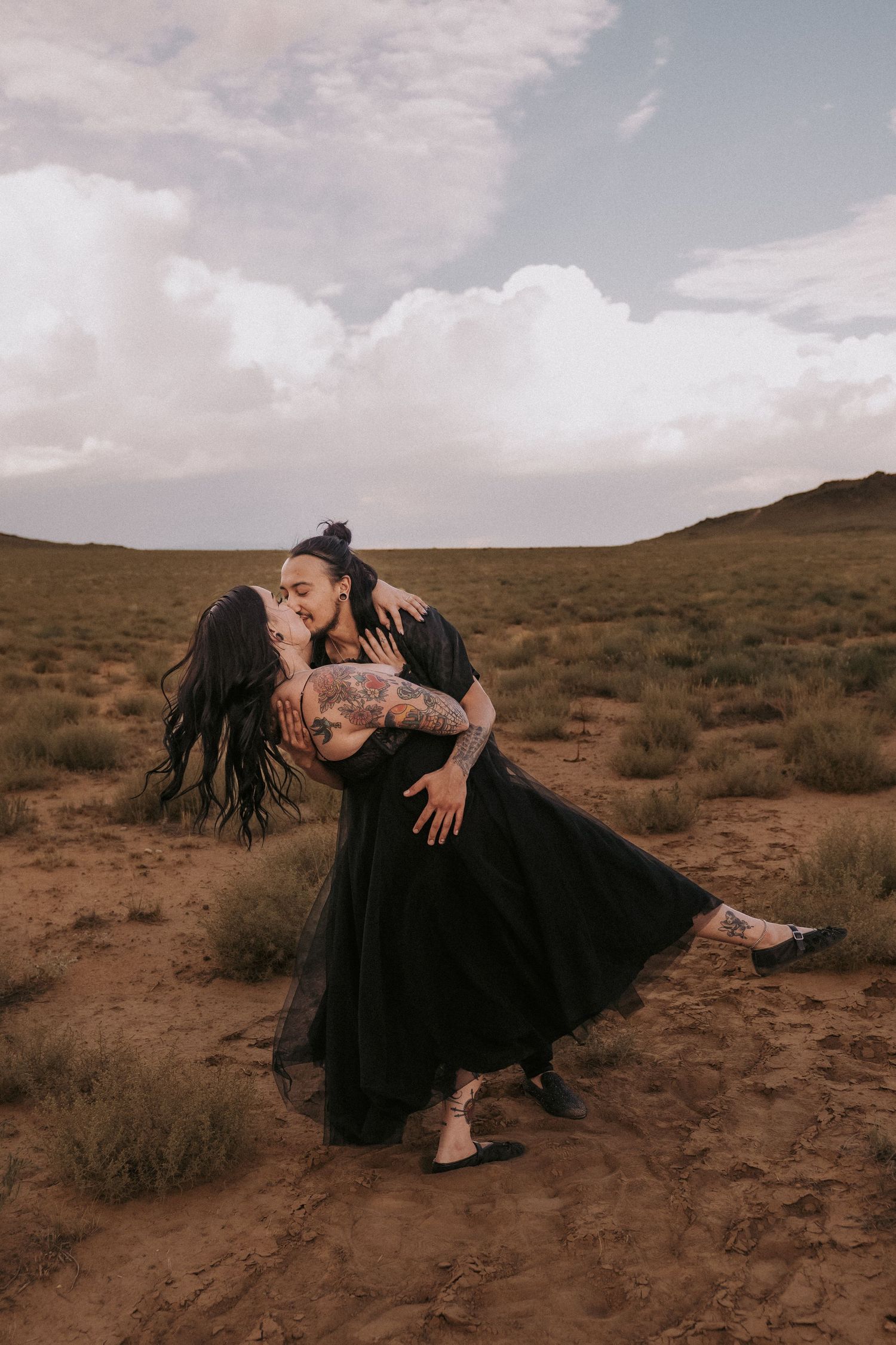 A romantic couple in black clothing share an intimate dance pose in a desert landscape near Albuquerque, NM under cloudy skies at sunset.