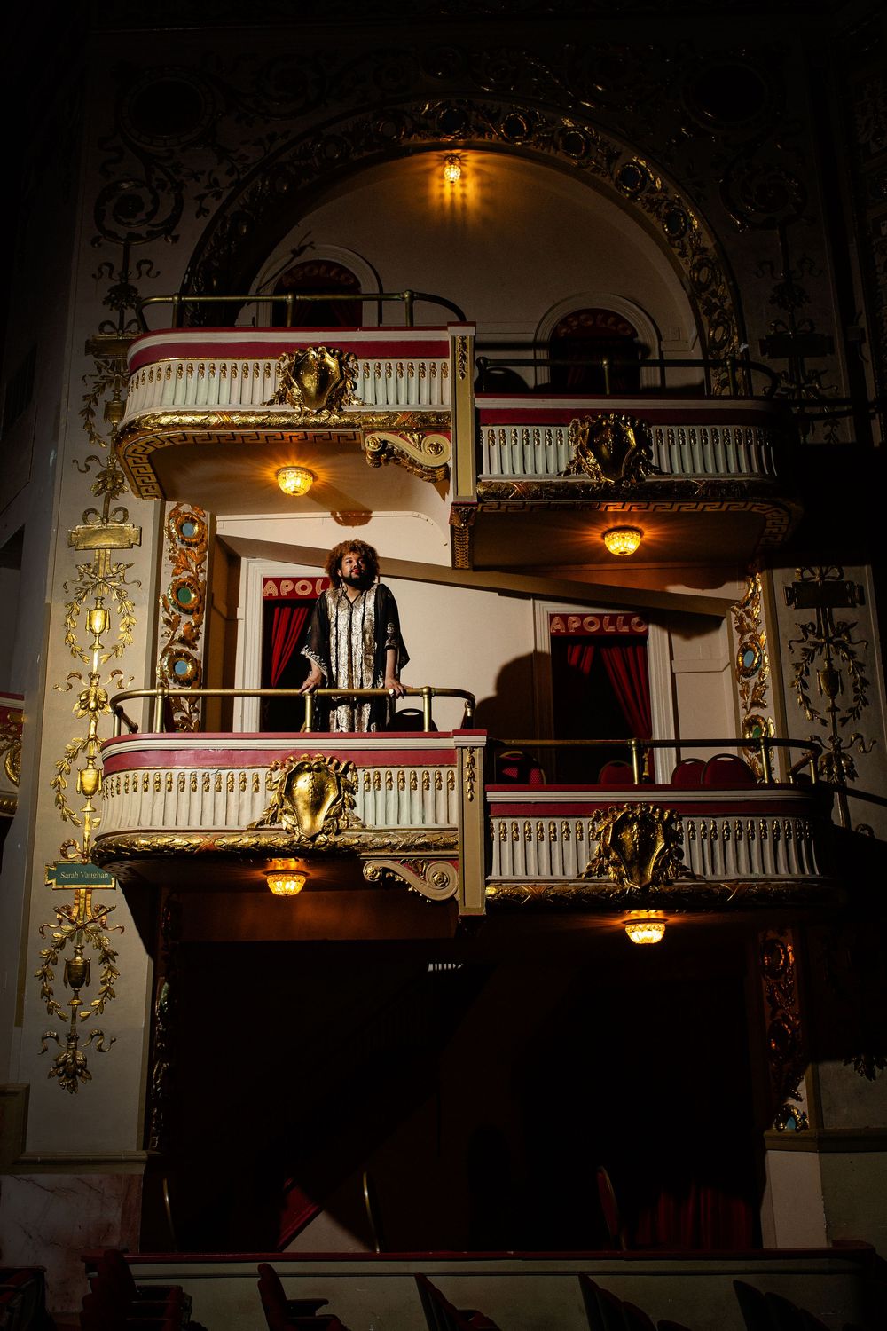Ornate Apollo Theater balcony with decorative gold trim, warm lighting, and a performer standing on the elegant tiered stage.