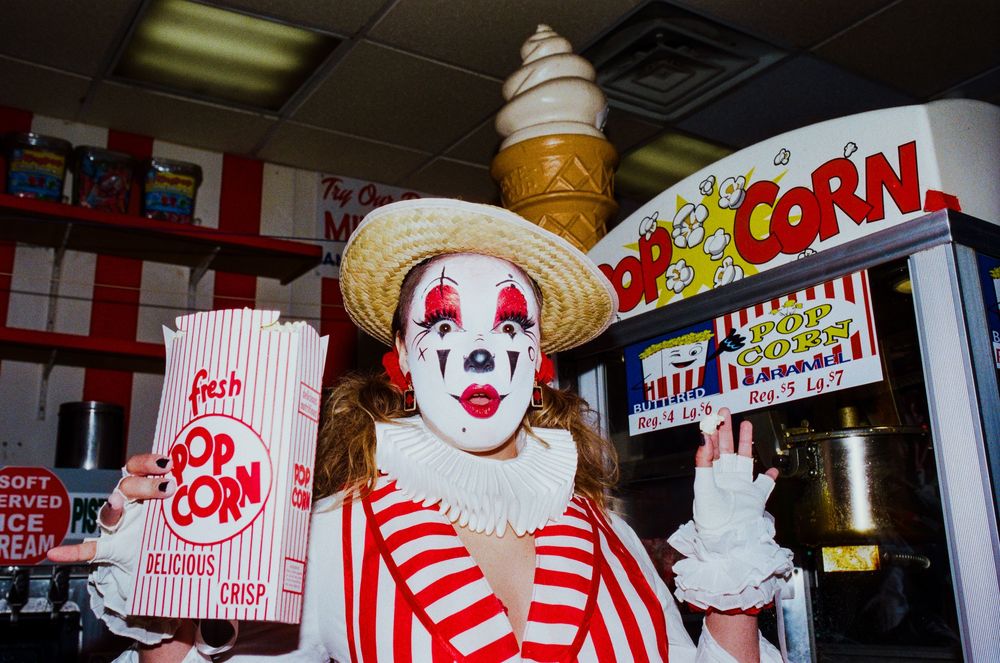A clown in a straw hat holds a popcorn bag near a soft serve ice cream machine and popcorn stand.