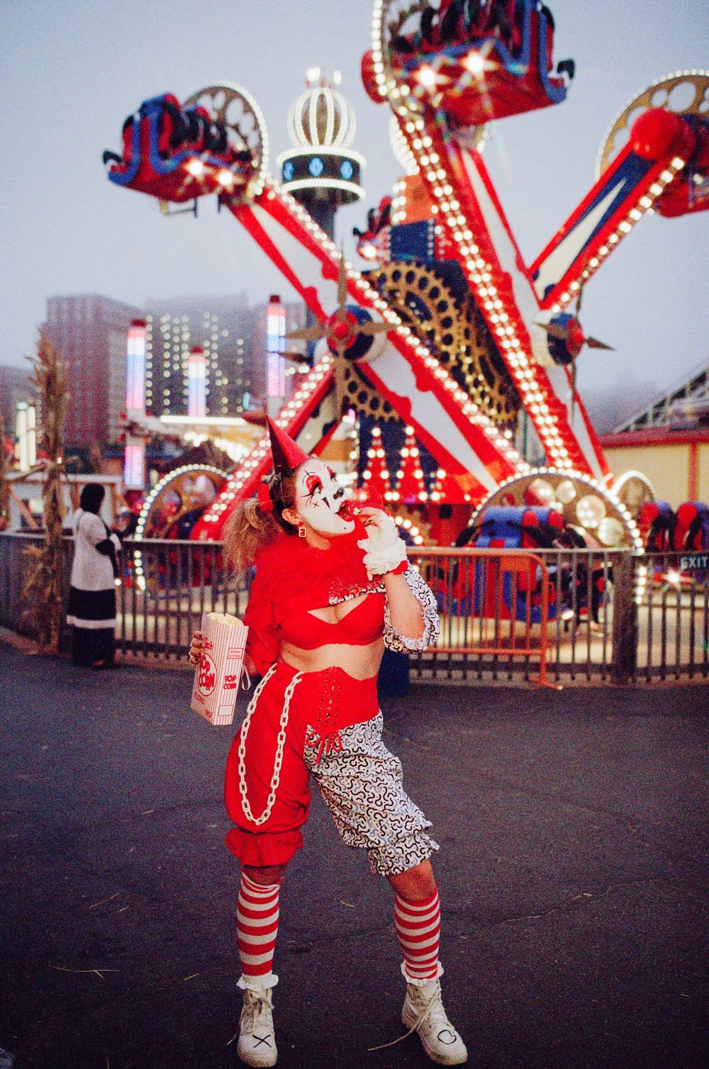 Person dressed as a clown at a colorful amusement park with bright rides and lights in the background.