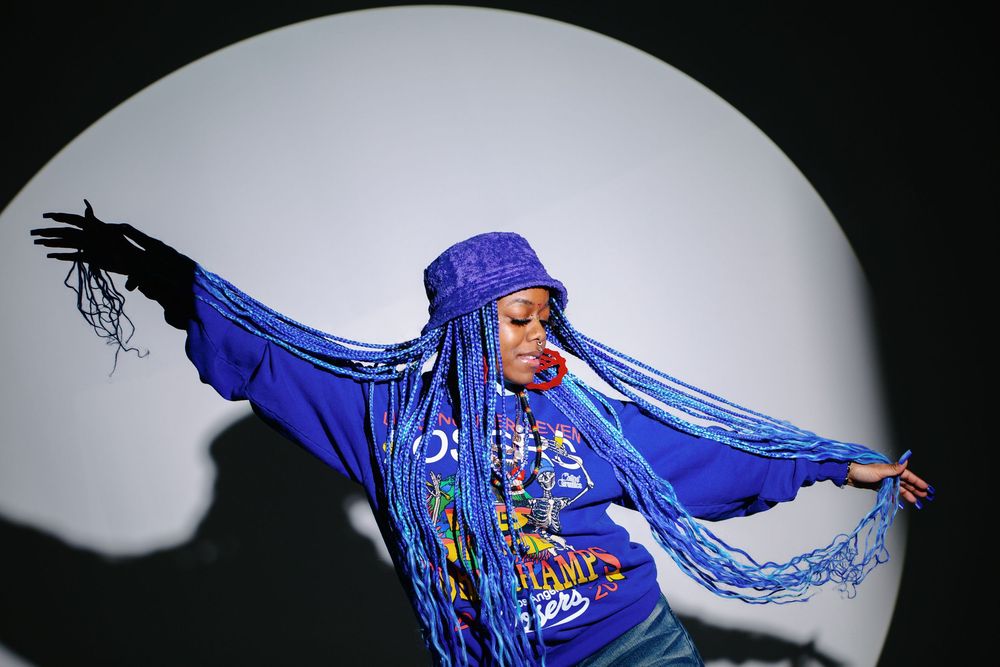 Person in blue hoodie and long blue braids poses dramatically with arms outstretched against circular backdrop.