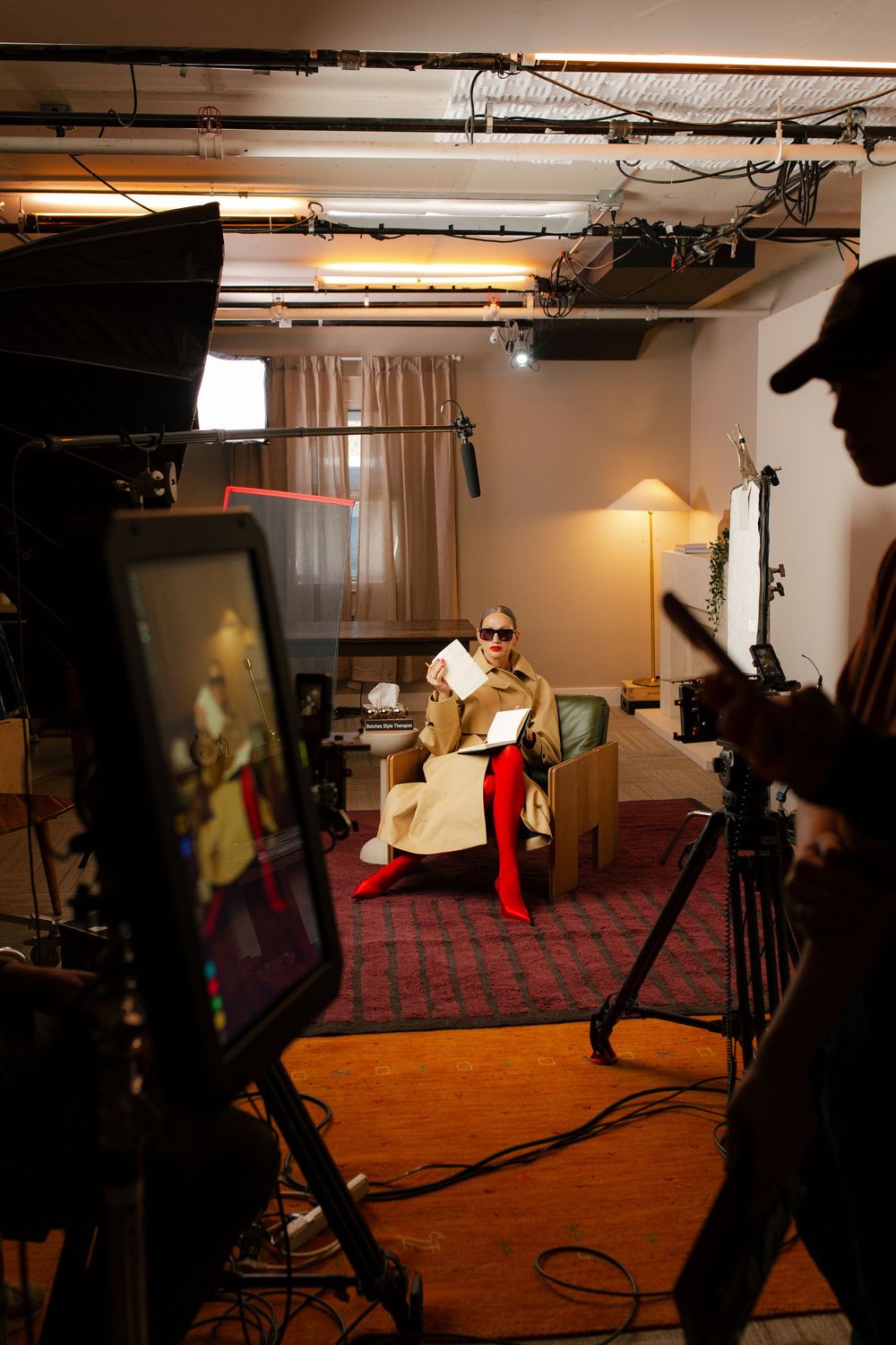 Behind-the-scenes film set with a person seated in a chair holding papers, surrounded by studio lighting and camera equipment.