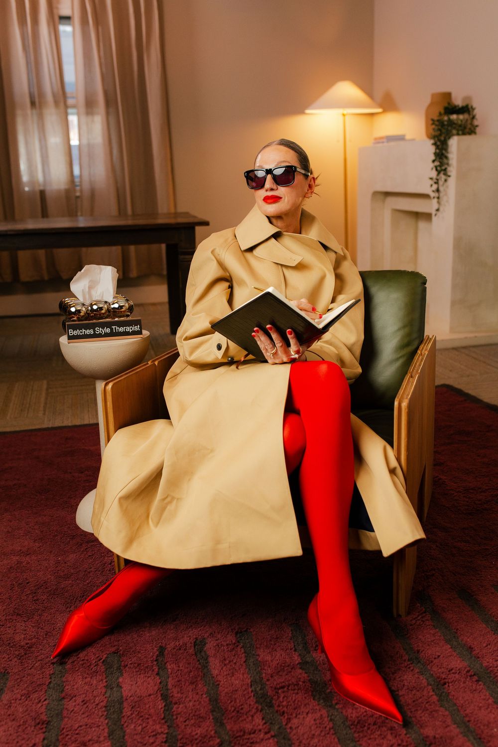 Stylish woman in beige trench coat and red thigh-high boots sits in armchair reading a book with sunglasses on.