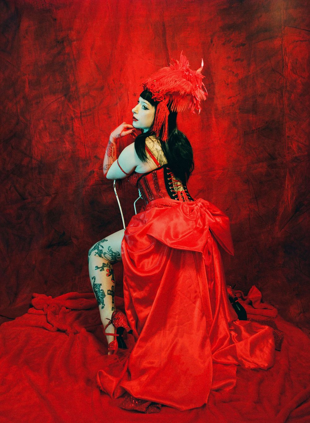 Woman in red corset gown with feathered headpiece poses dramatically against a rich red backdrop in burlesque style.