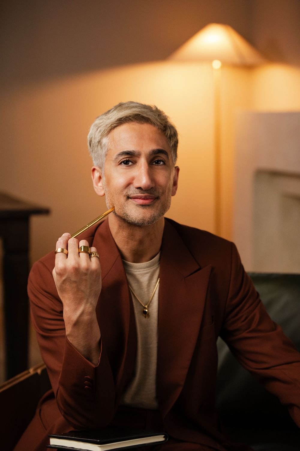 Stylish man in brown blazer holding gold pencil, wearing rings and necklace, seated near warm lamp light.
