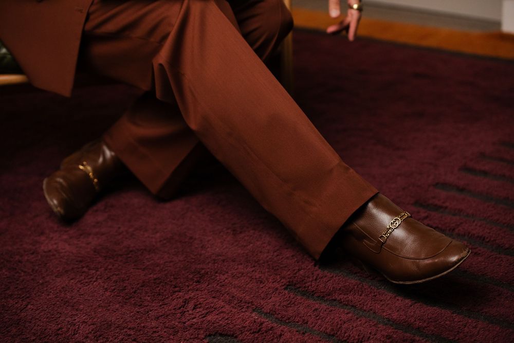 Man in brown trousers and leather loafers seated on a burgundy carpet, showcasing stylish monochromatic fashion.