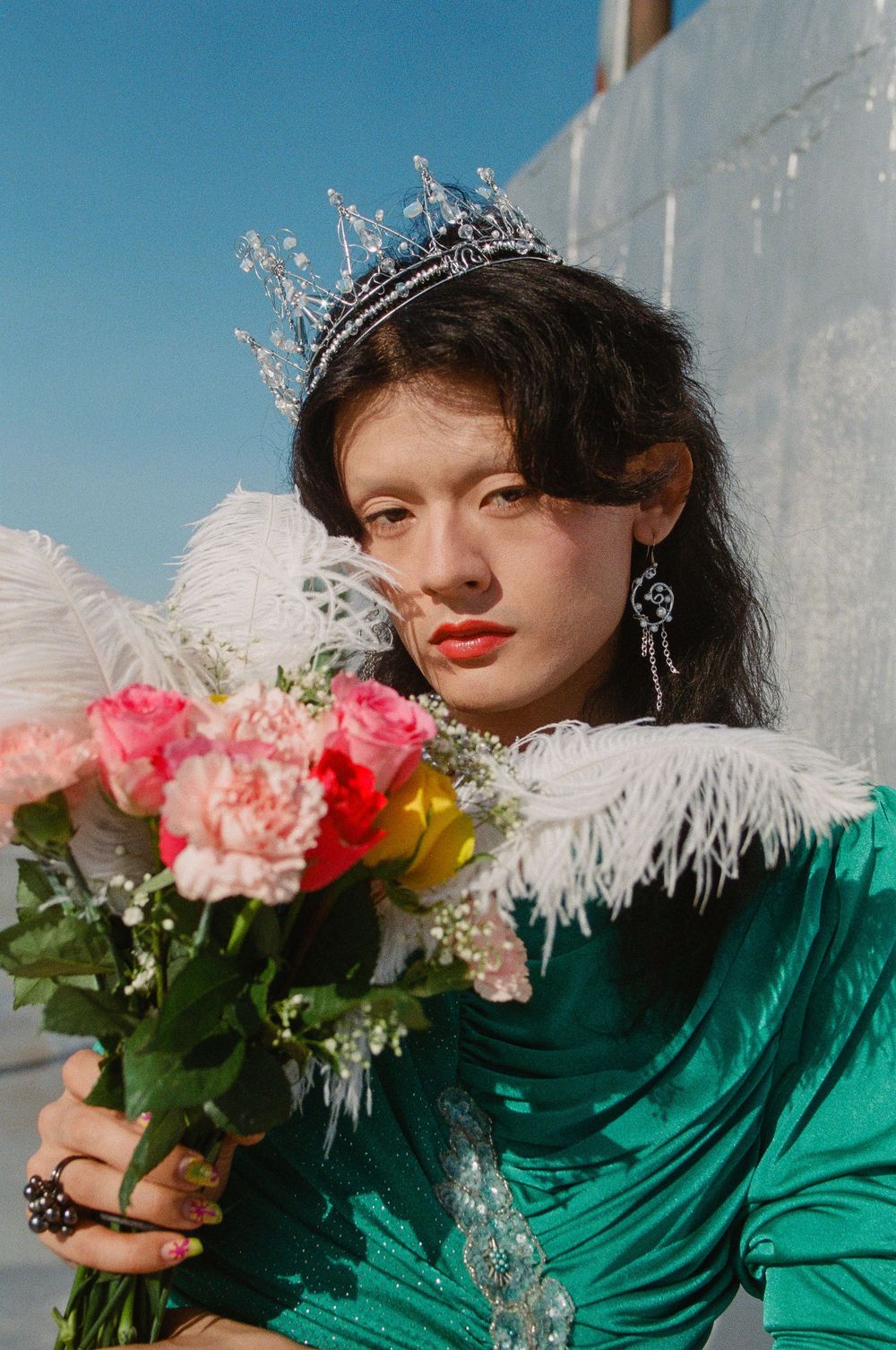 Elegant portrait featuring a crystal crown, emerald velvet dress, and pink floral bouquet against a bright blue sky.