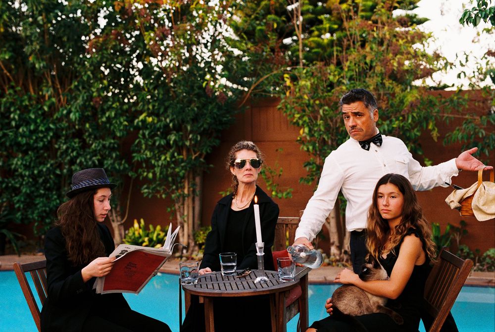 Elegant group at poolside table with waiter in bow tie, two women seated, one reading menu outdoors.