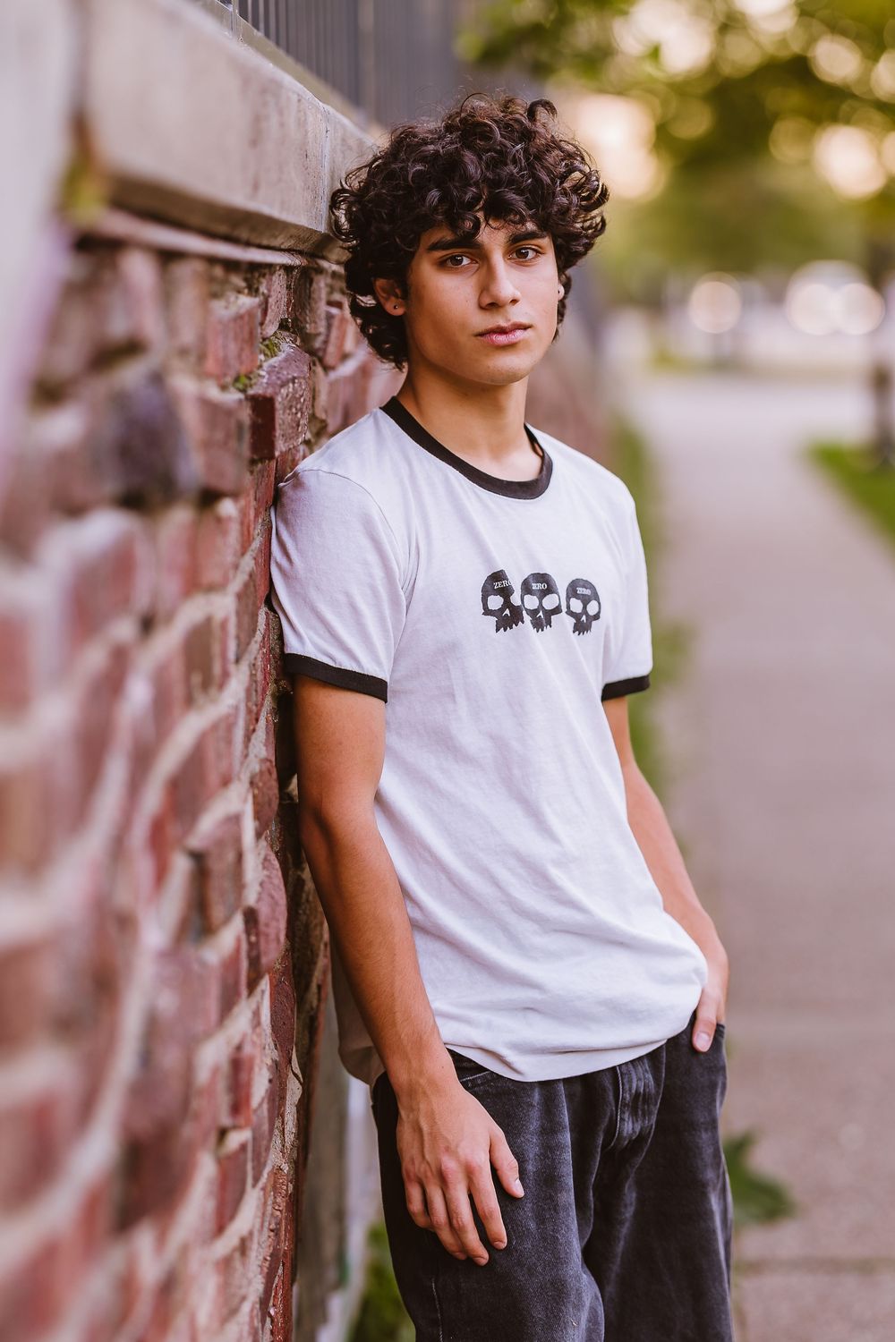 A person in a white ringer t-shirt with skull graphics leans against a brick wall in warm evening light.