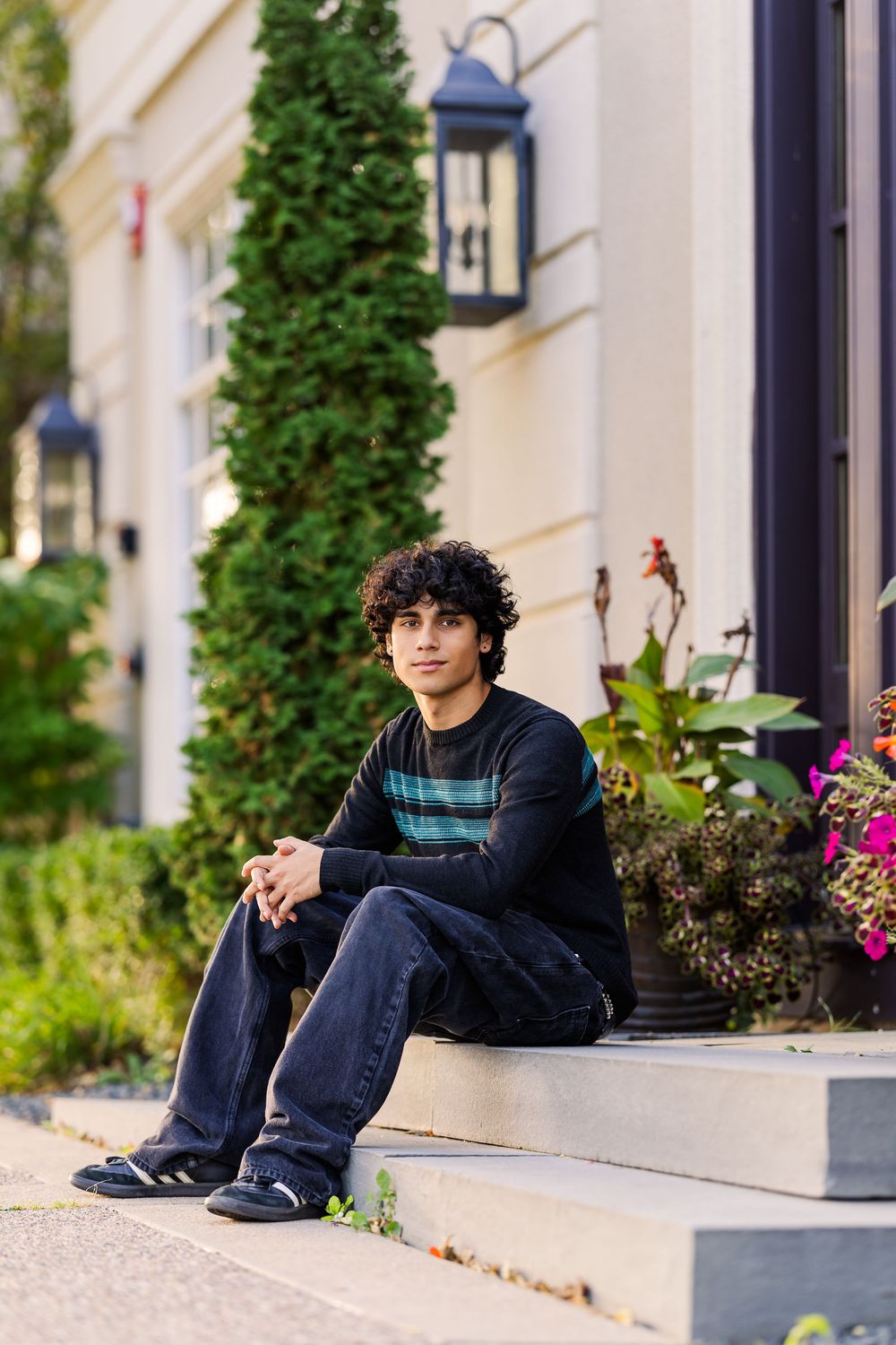 A person sits on outdoor steps wearing a striped sweater and jeans, with greenery and a lantern visible on the building.