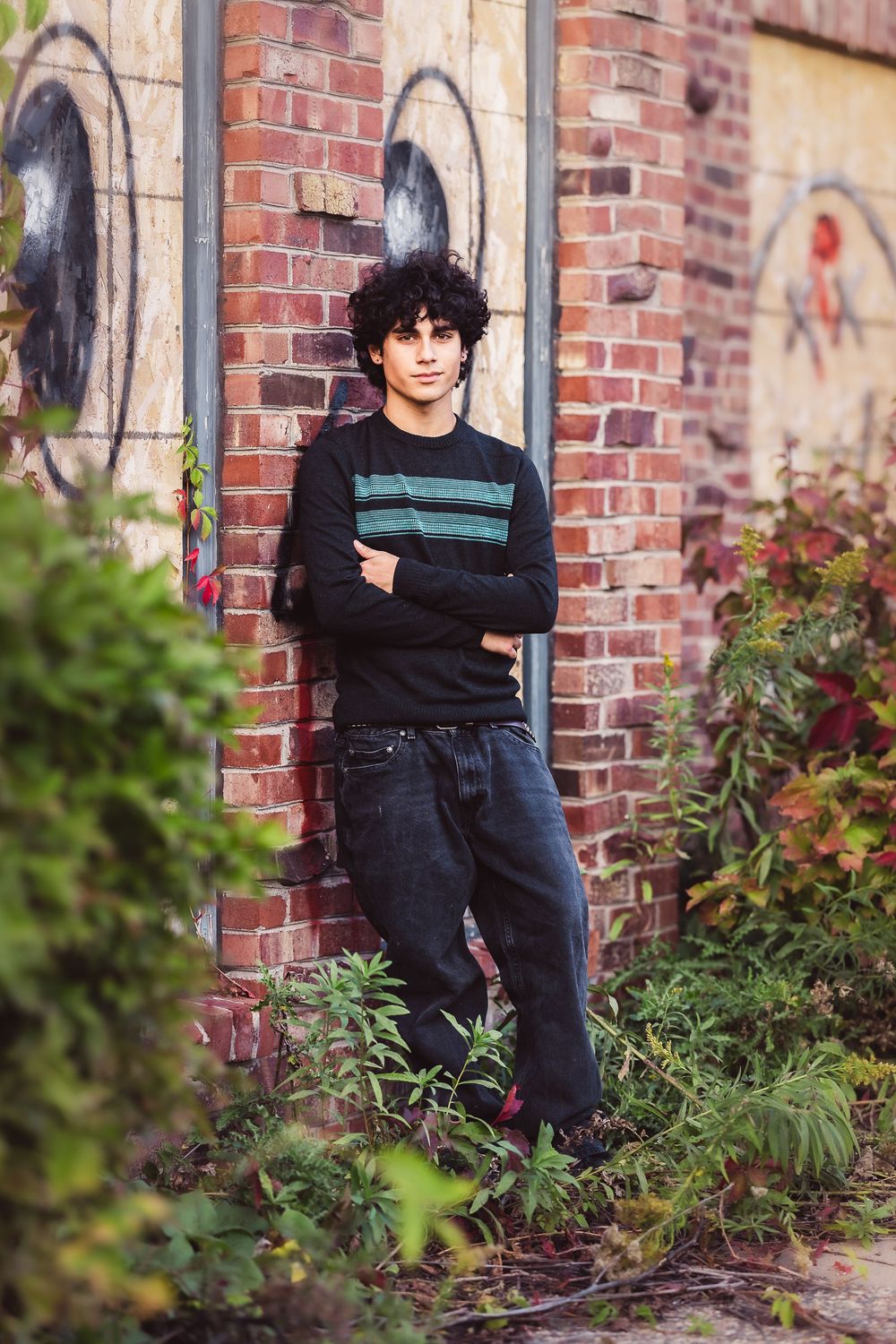 Person in dark clothing sits against a brick wall surrounded by garden foliage.