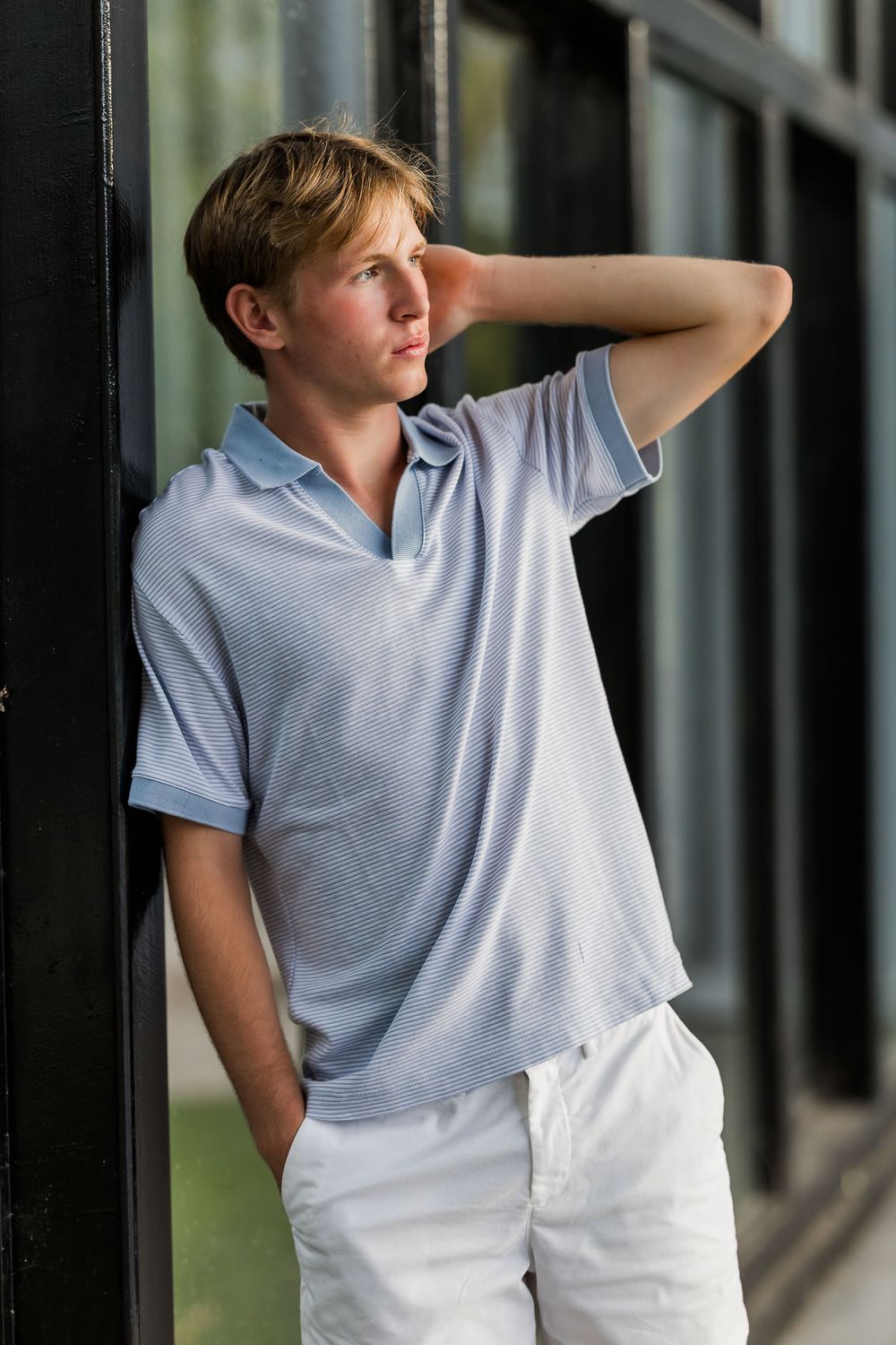 A young person in a light gray polo shirt and white pants poses casually against a dark background.