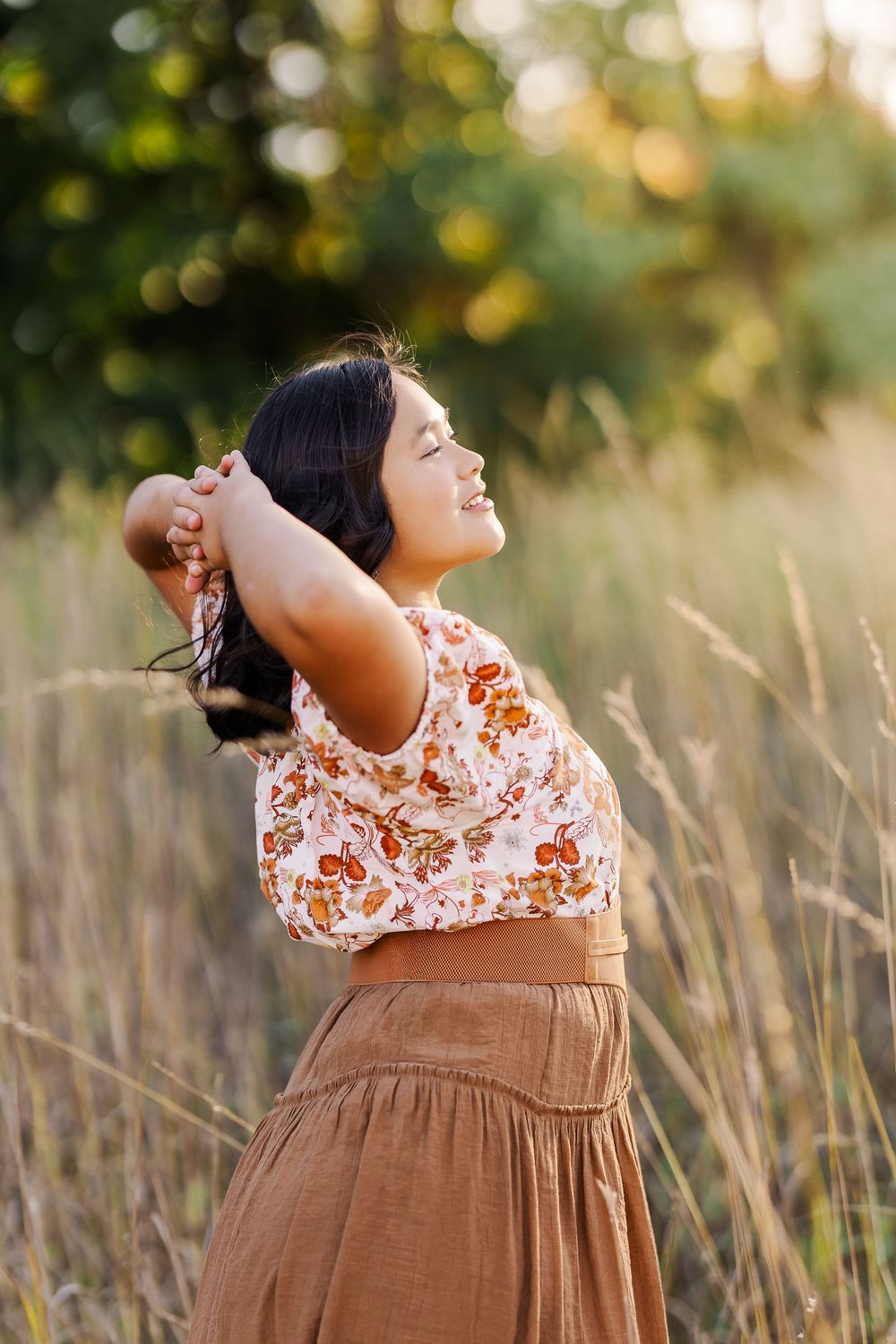A person in a floral top and brown dress stands in tall grass at golden hour with their head tilted back.