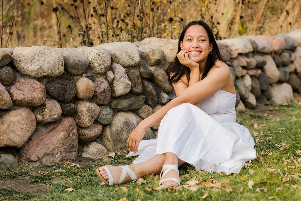 Person in white dress sitting by stone wall during fall season.