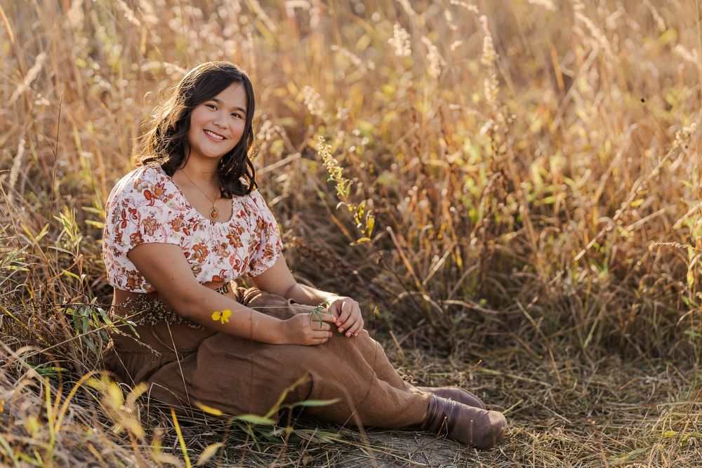 A person in a floral dress sits in a golden field at sunset wearing brown boots and smiling warmly.