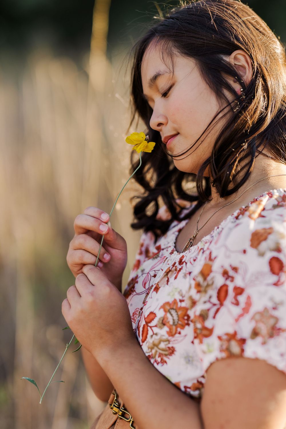 A child in a floral dress smells a small yellow flower in a series of dreamy outdoor portraits at golden hour.
