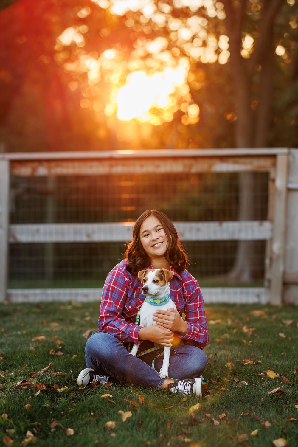 A person in plaid shirt sits on autumn grass with small white dog at golden sunset behind white fence.