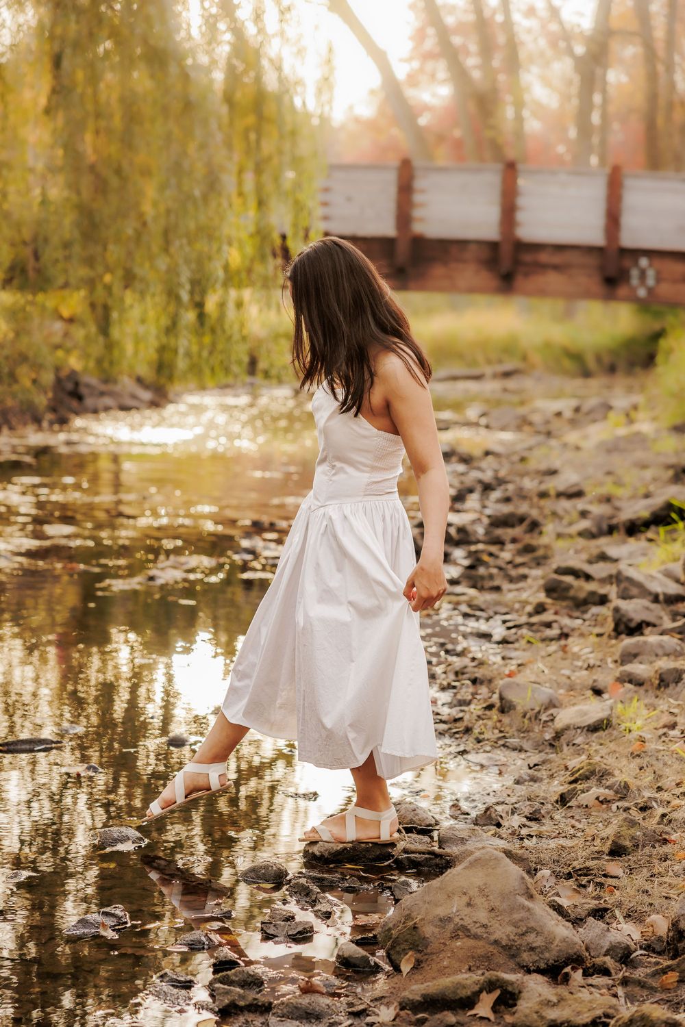 Young woman in white dress standing by stream edge exploring nature on sunny day.