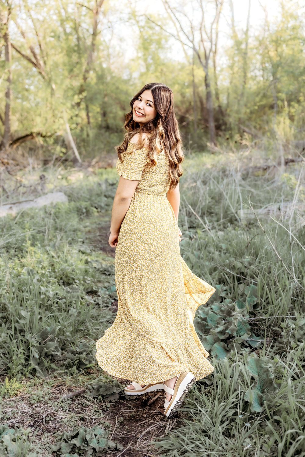 Person wearing a flowing yellow maxi dress poses in a natural outdoor setting with tall grass and trees in background.