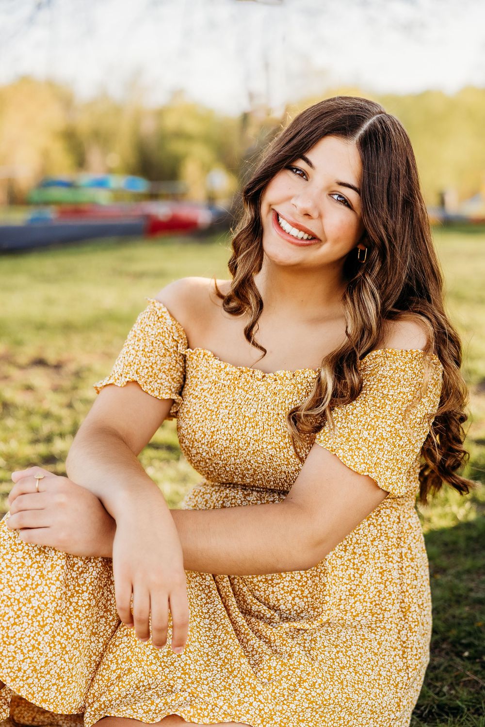Smiling person in yellow floral off-shoulder dress poses outdoors during golden hour in parklike setting.