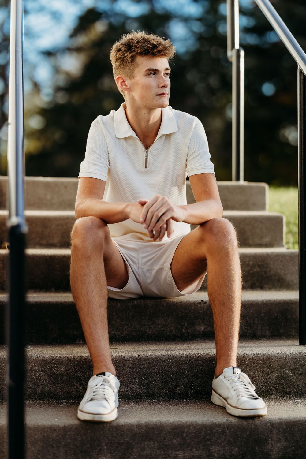 Person in white polo shirt and shorts sitting on concrete steps wearing white sneakers in outdoor setting.