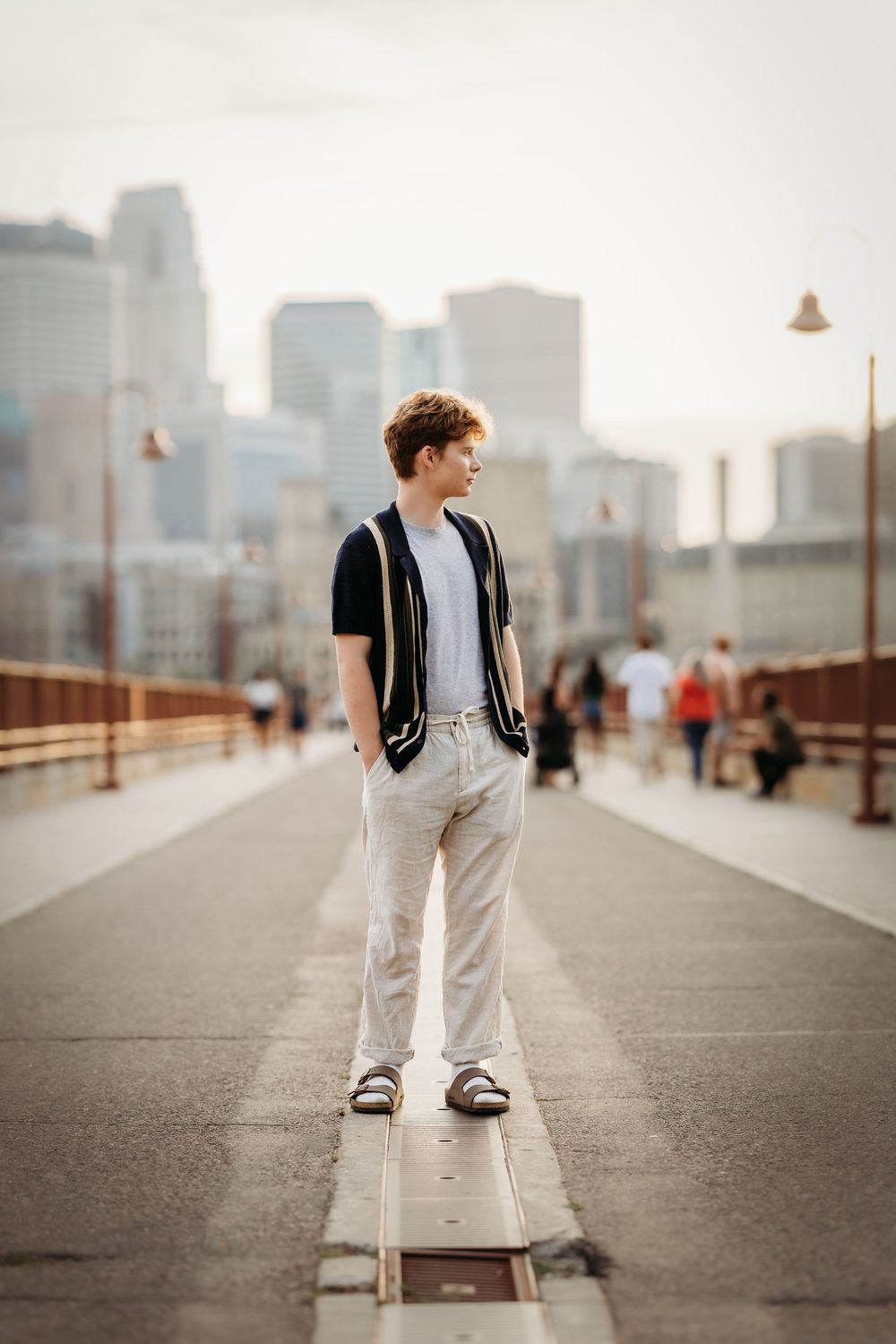 Person in black vest and white pants stands on urban boardwalk with city skyline in background.