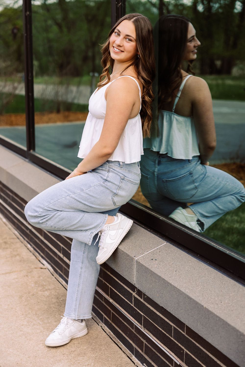 A casual summer look with someone wearing a white tank top, light wash jeans and white sneakers sitting on a reflective ledge.