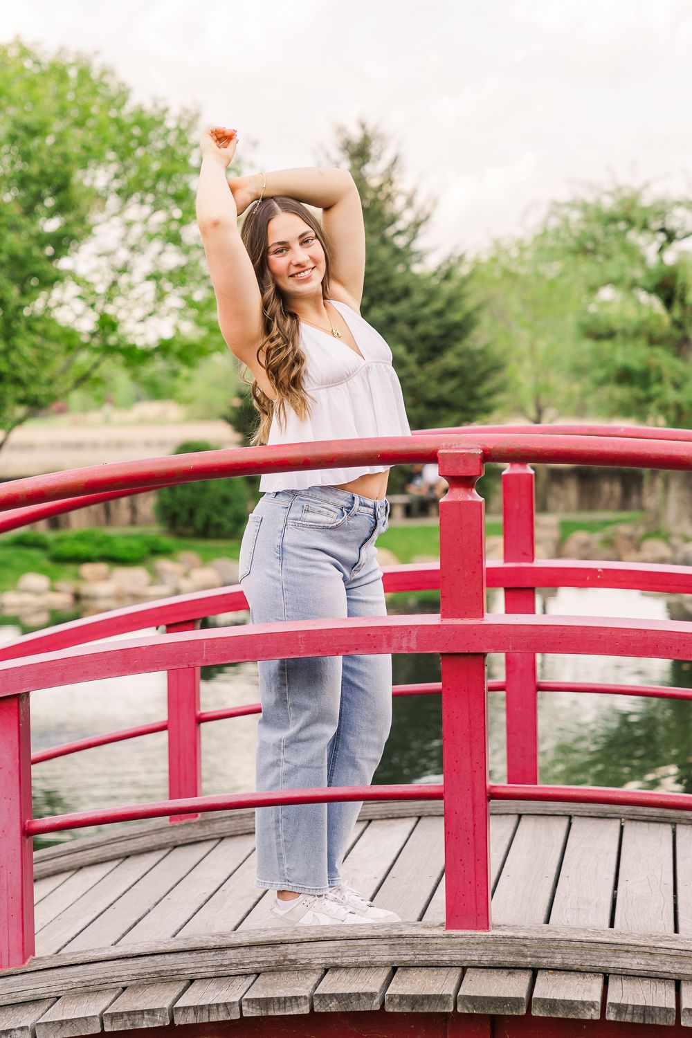Woman in white top and jeans poses joyfully on a curved red Japanese garden bridge surrounded by green foliage.