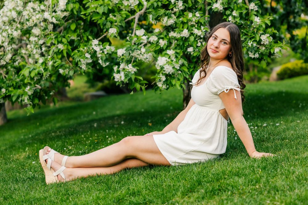 Person in white sundress sits on grass beneath blooming apple trees in spring garden.