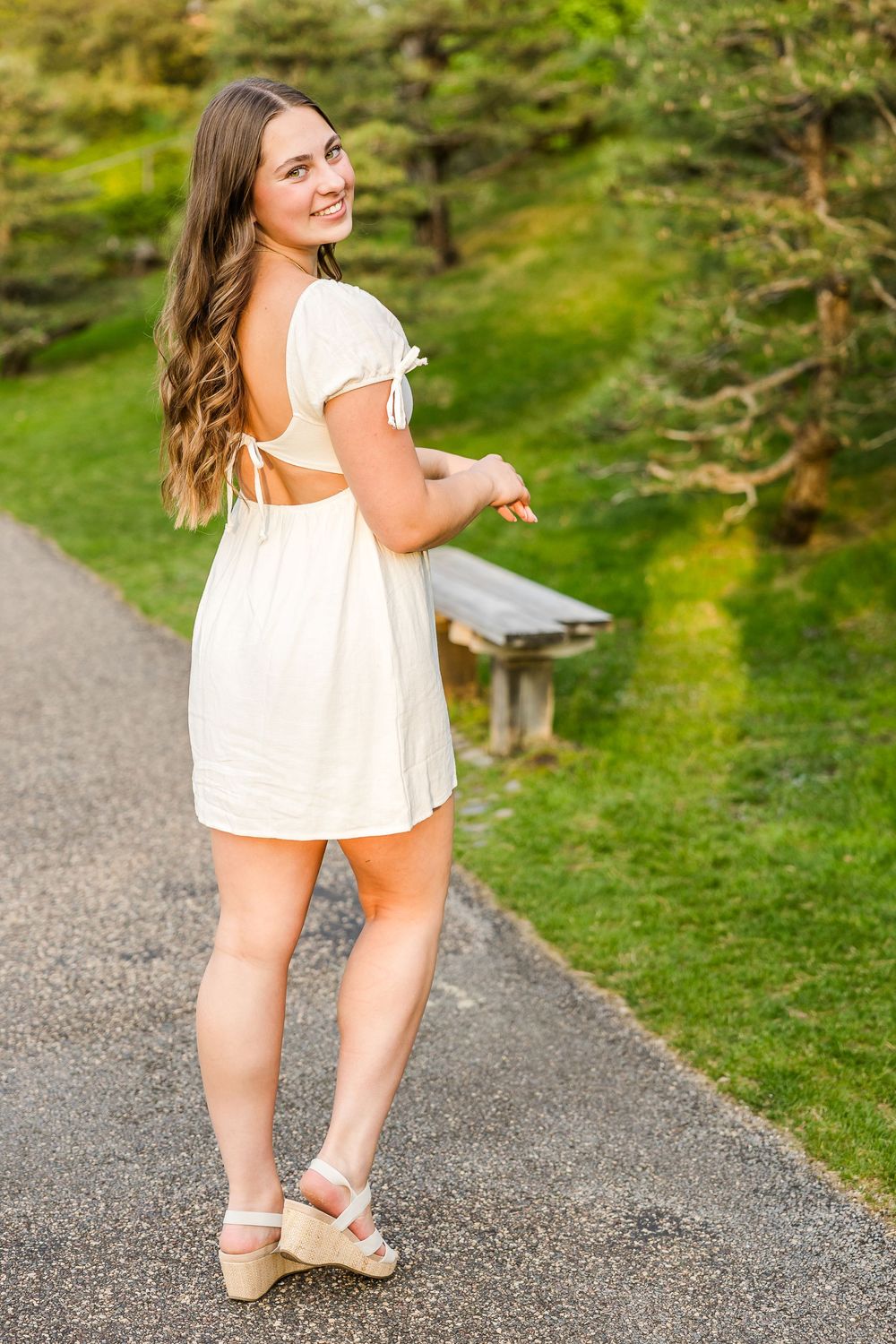 Looking back while wearing a white summer dress and wedge sandals on a path in a lush green outdoor setting.