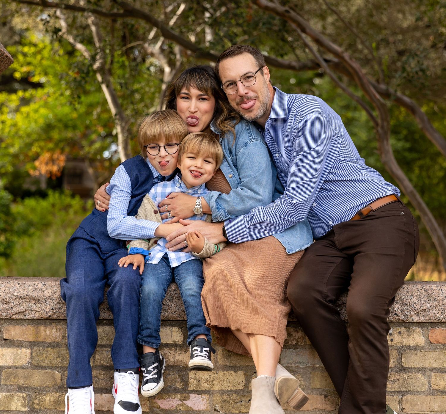 Happy family sitting together on stone wall in outdoor garden setting with greenery in background.