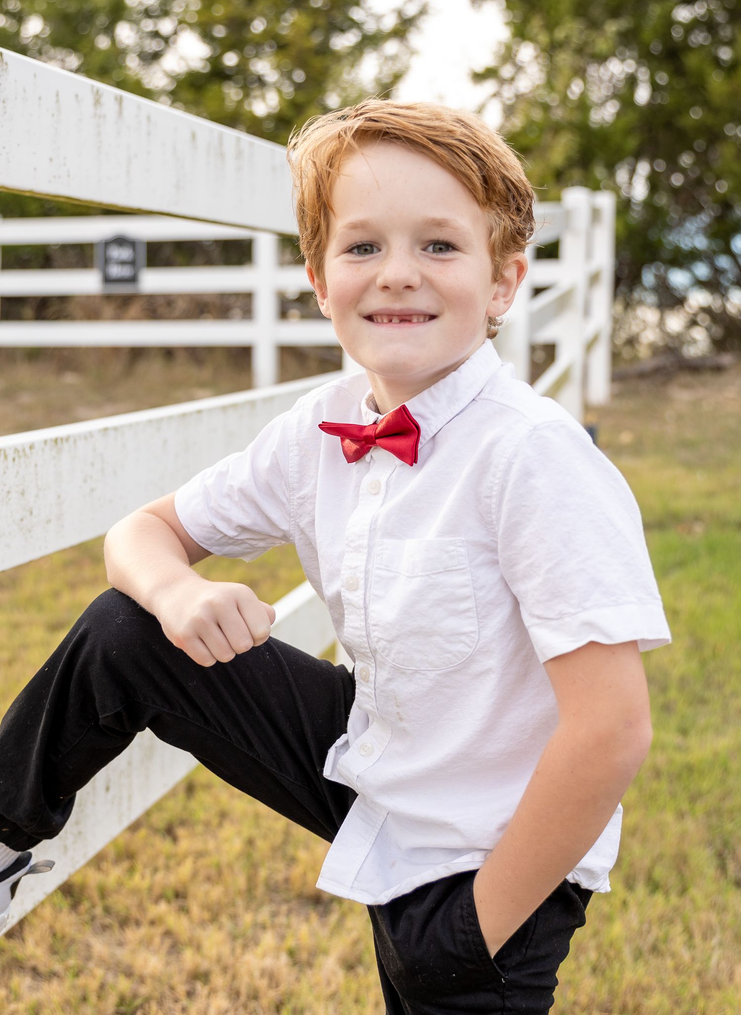 Young child in white shirt and red bowtie poses casually by white fence in outdoor setting.