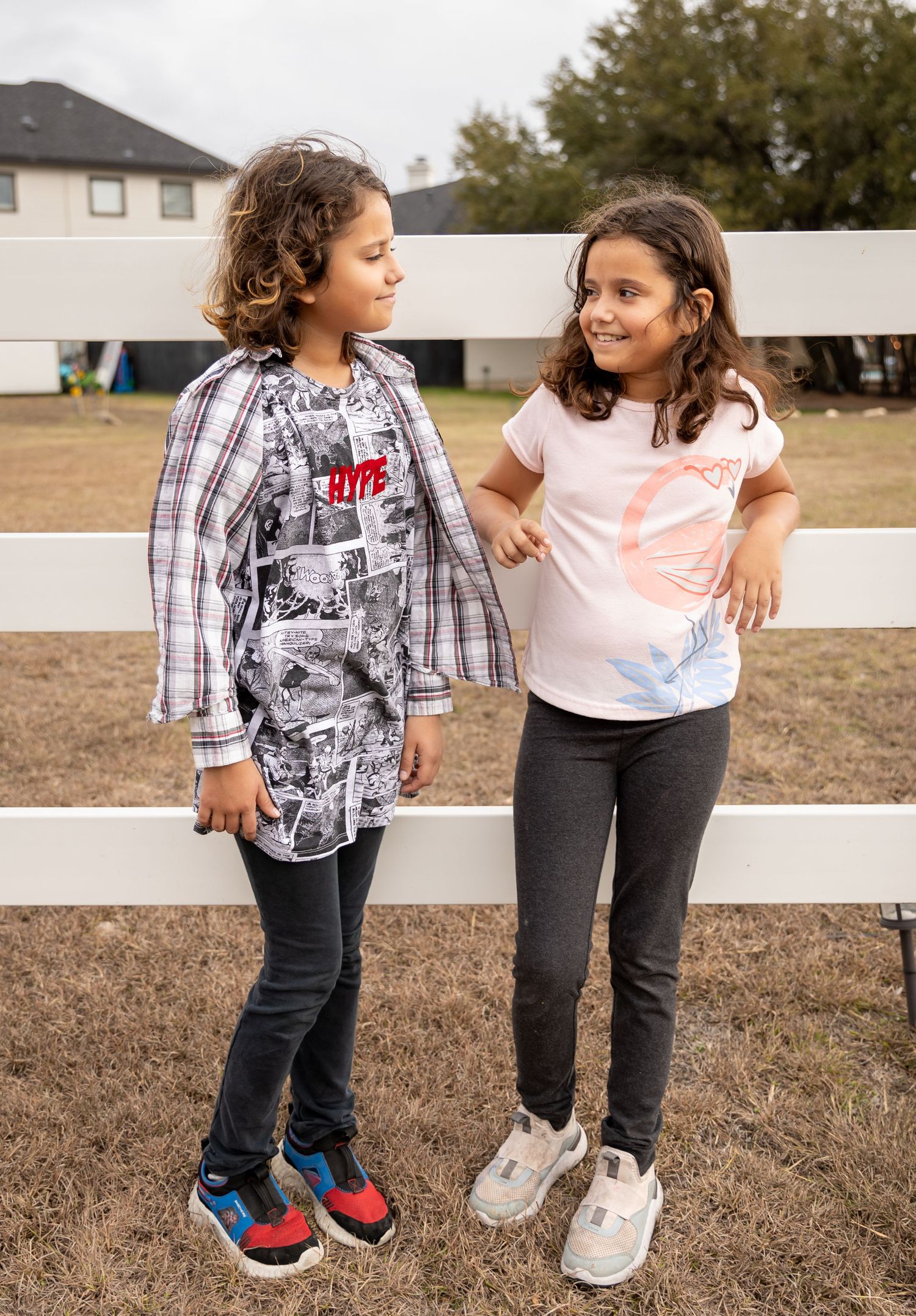 Two children in casual attire chat while standing by a white fence outdoors.