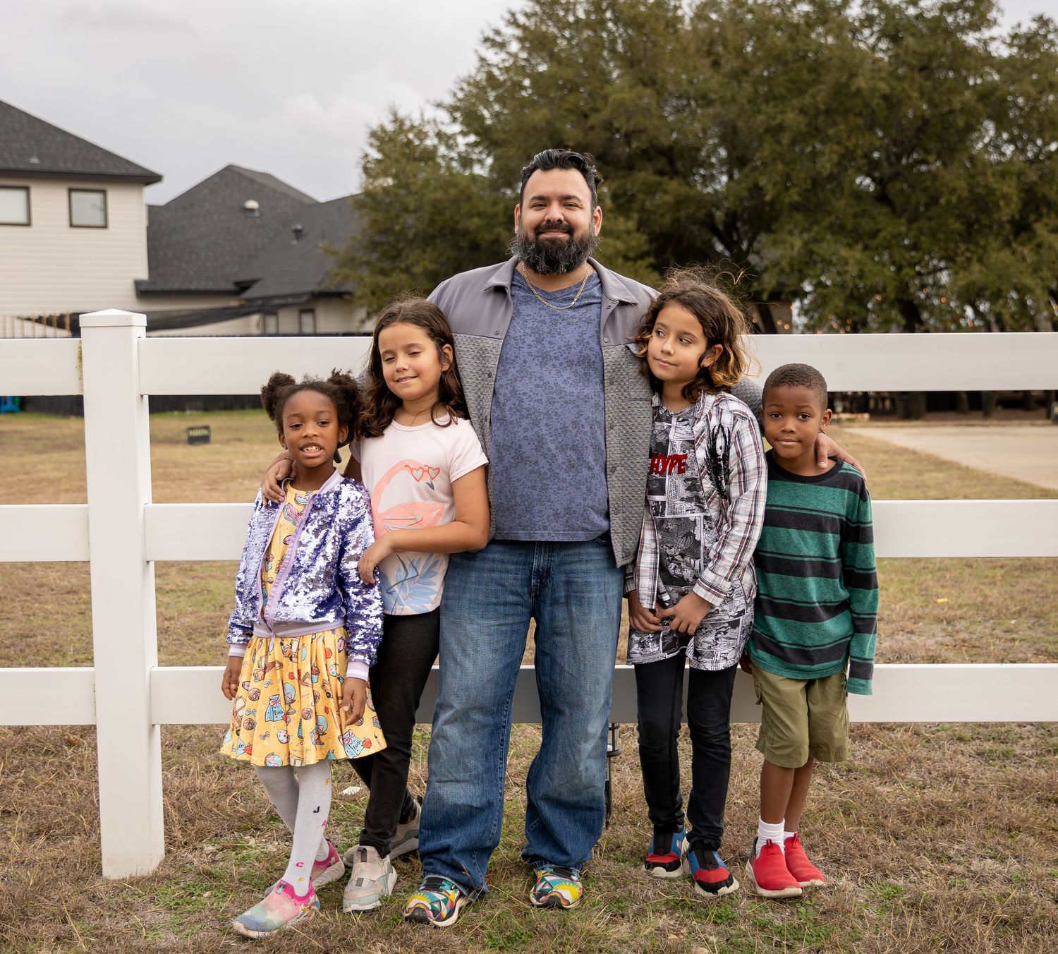 A family stands together in front of a white fence on a residential property.