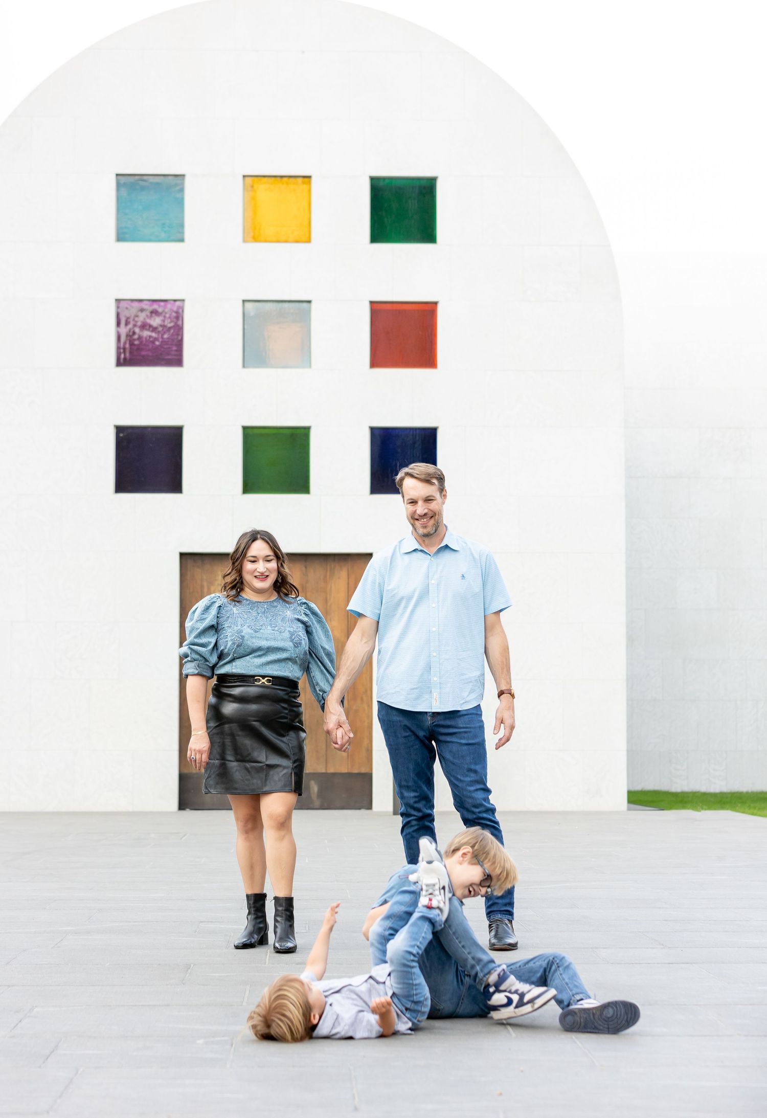 Family poses against white wall with colorful squares as children play on ground.