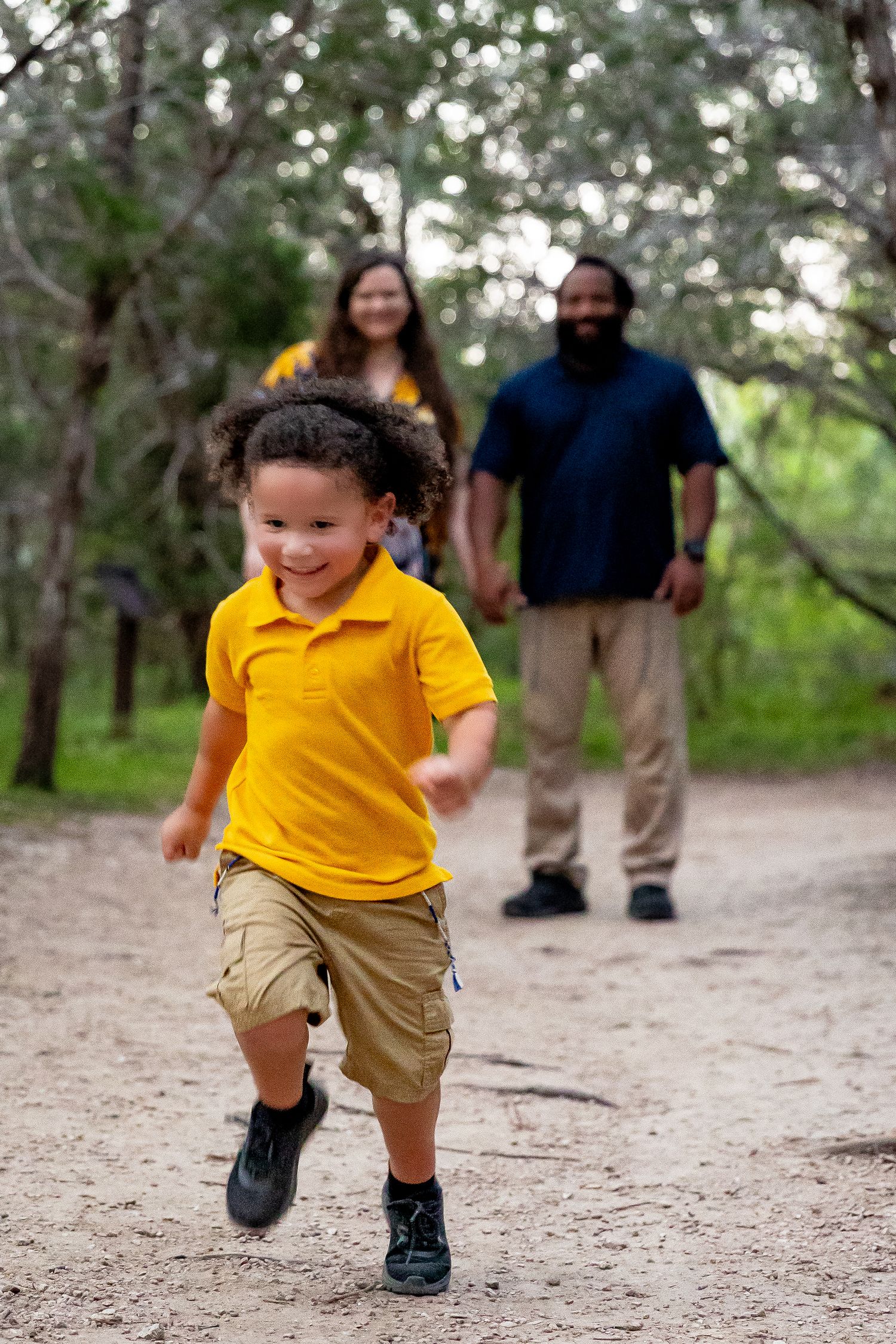 A joyful child in a yellow polo shirt runs along a nature trail while family members follow behind.