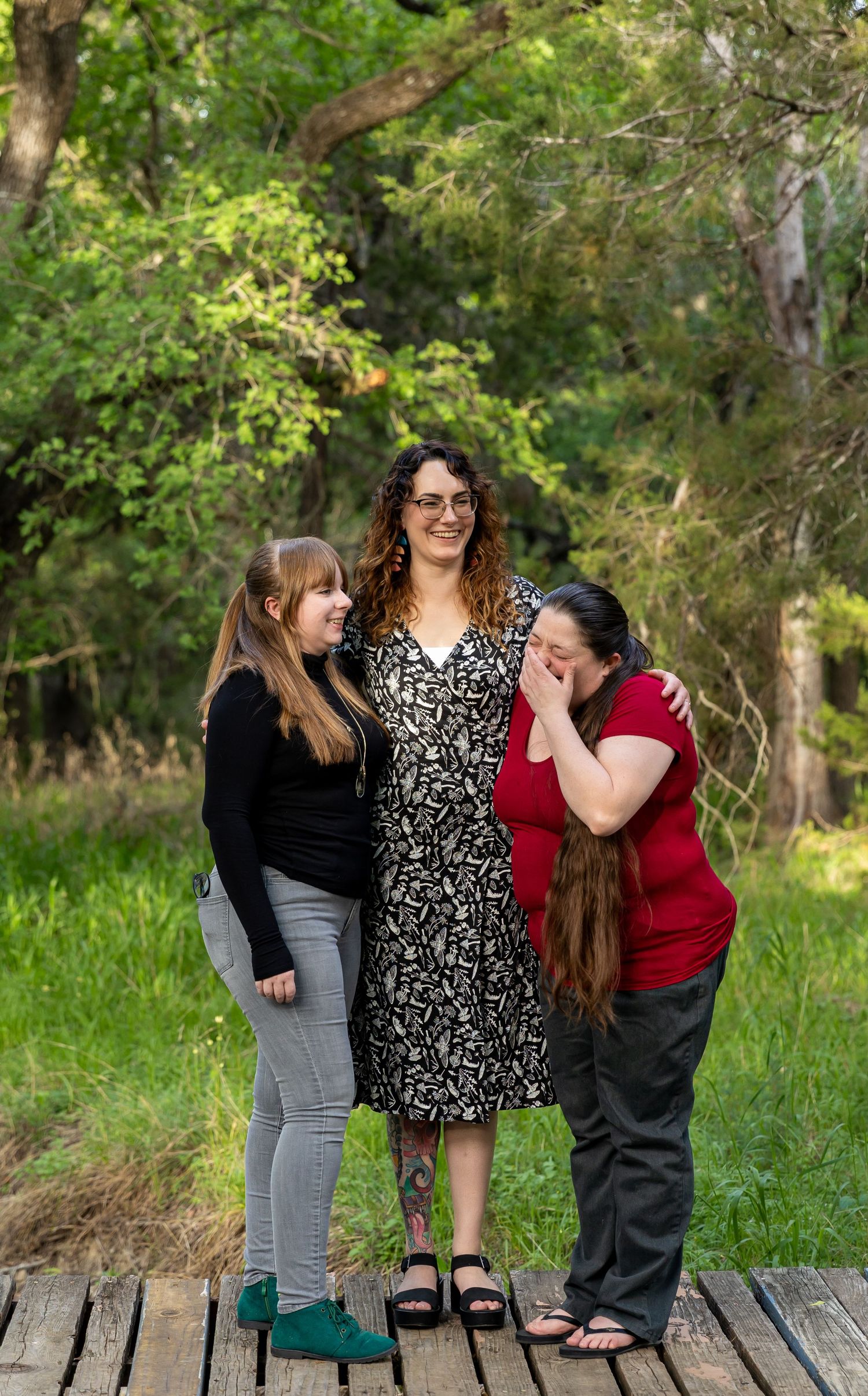 Three people share a lighthearted moment while standing on a wooden deck in a lush forest setting.
